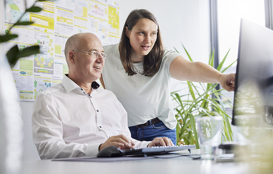 Mann sitzt vor PC, Frau steht daneben und zeigt auf den Bildschirm