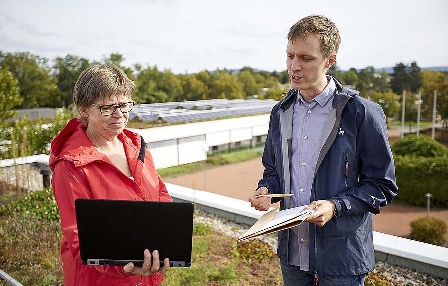 Frau und Mann stehen auf einem Schuldach mit Photovoltaik und schauen in einen Laptop