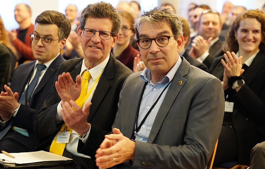 Drei Männer im Vordergrund vor größerem Publikum klatschen